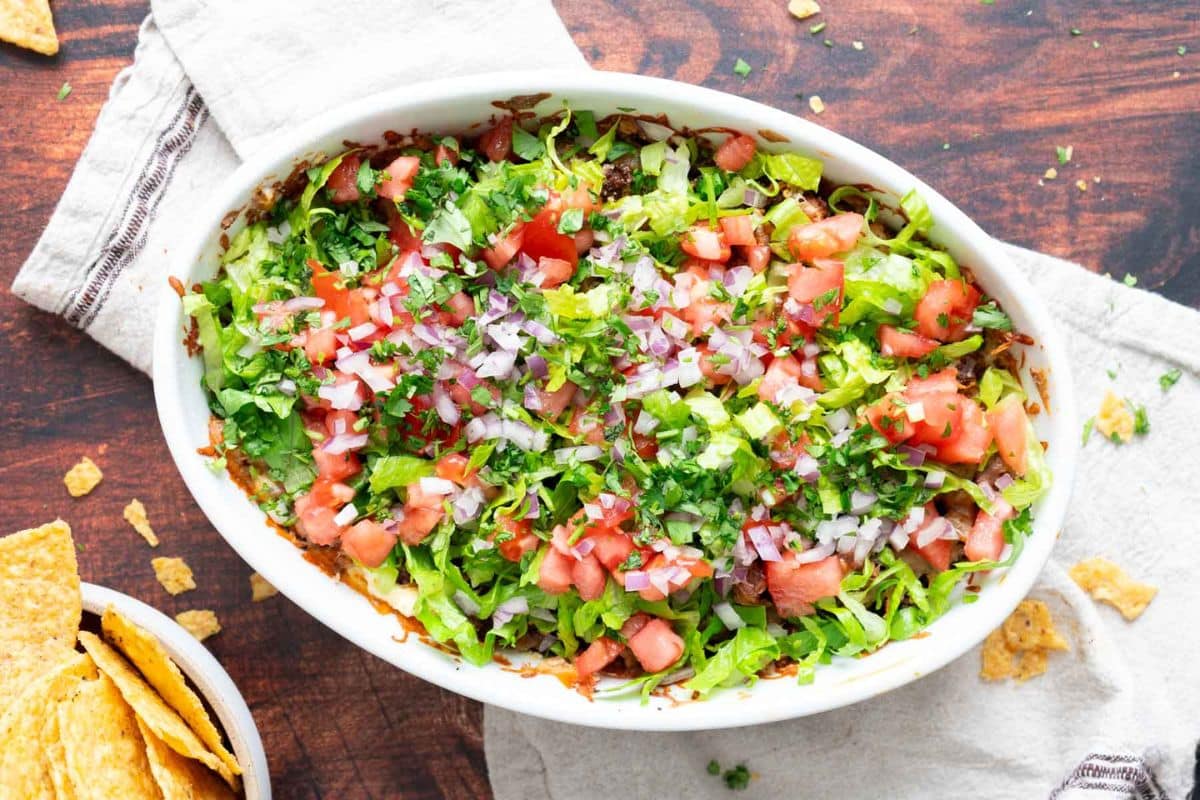 An oval dish filled with layered dip topped with chopped lettuce, tomatoes, red onions, and cilantro-perfect for Game Day Dips-with crunchy tortilla chips on the side.