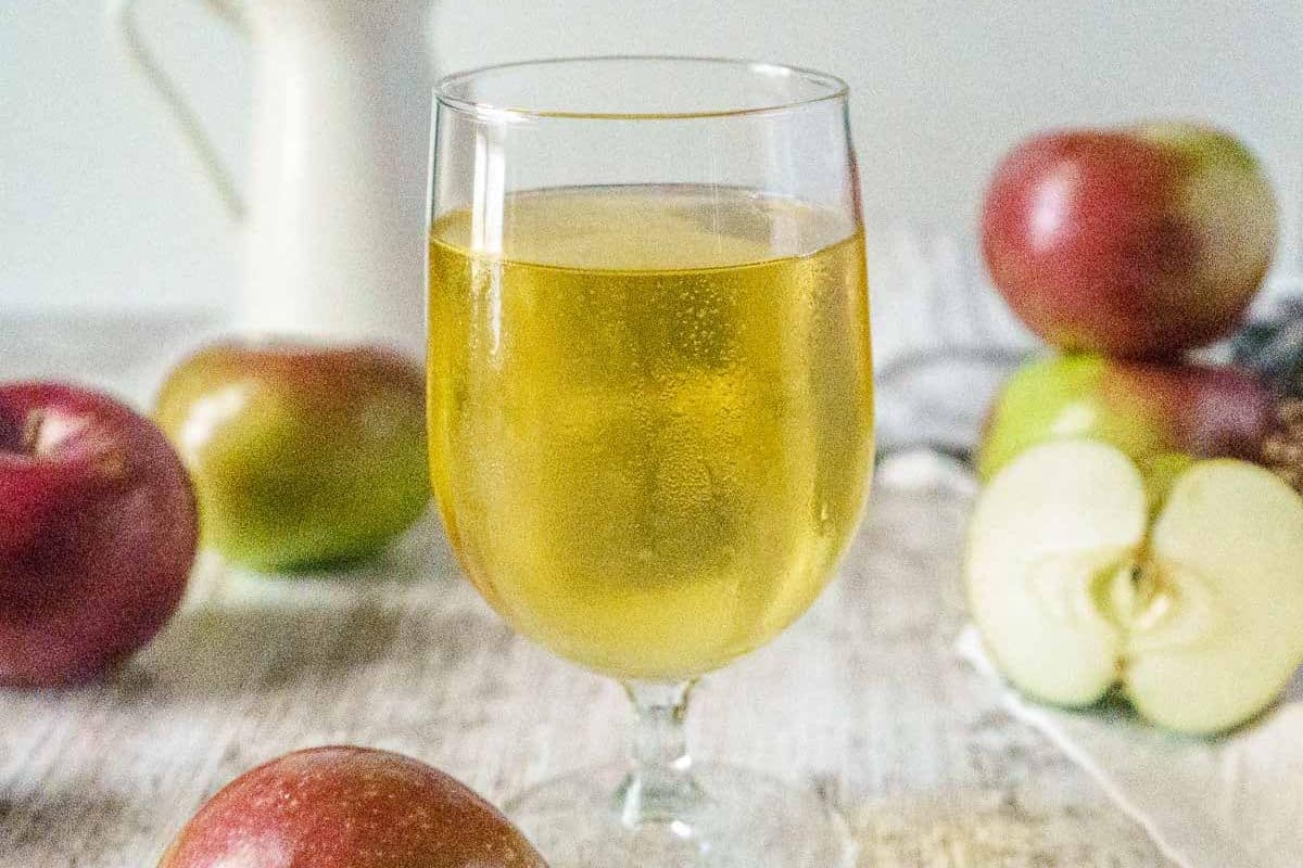 A glass of apple cider sits on a wooden surface surrounded by whole and sliced apples, inspiring sweet apple ideas and delicious apple recipes.