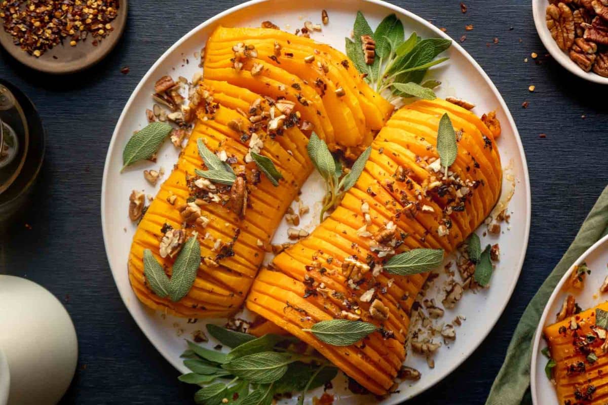 Three roasted and sliced butternut squashes garnished with chopped pecans, sage leaves, and red pepper flakes on a white plate-a standout among squash dishes perfect for any gathering.