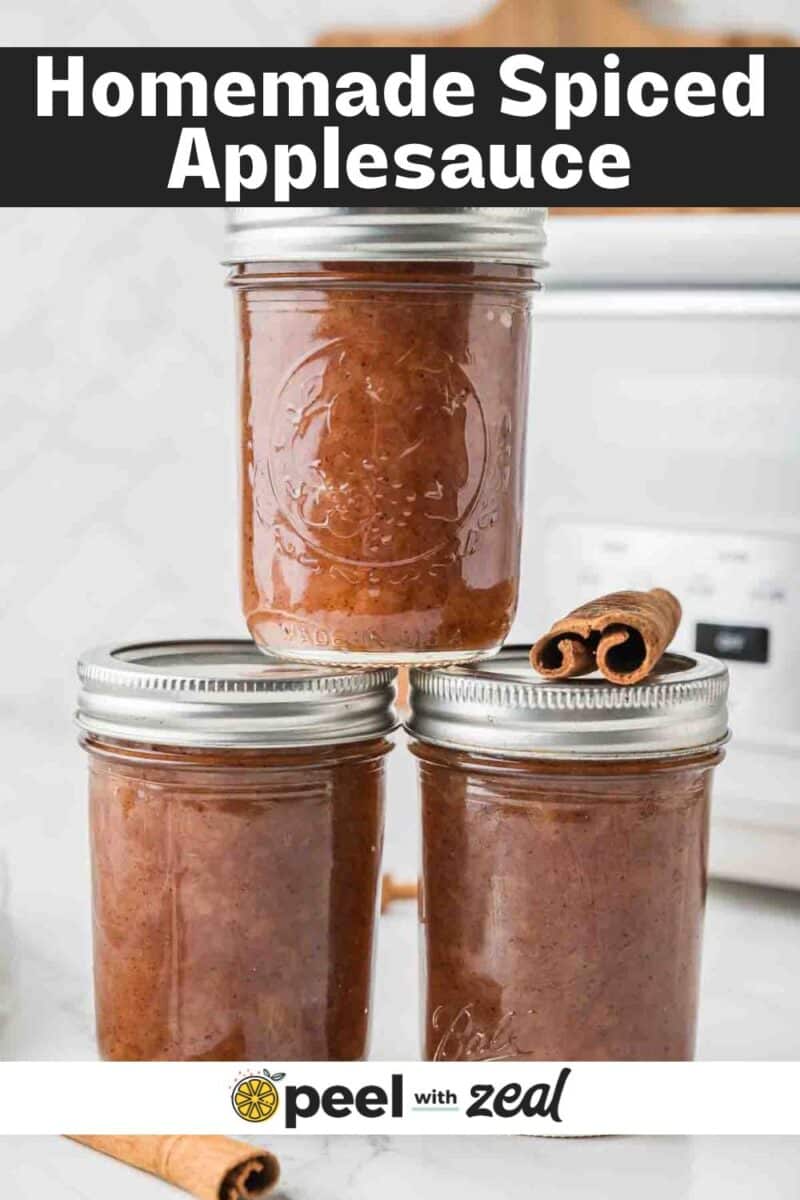 Three glass jars filled with homemade slow cooker applesauce sit on a countertop, with two cinnamon sticks placed on top of one jar.