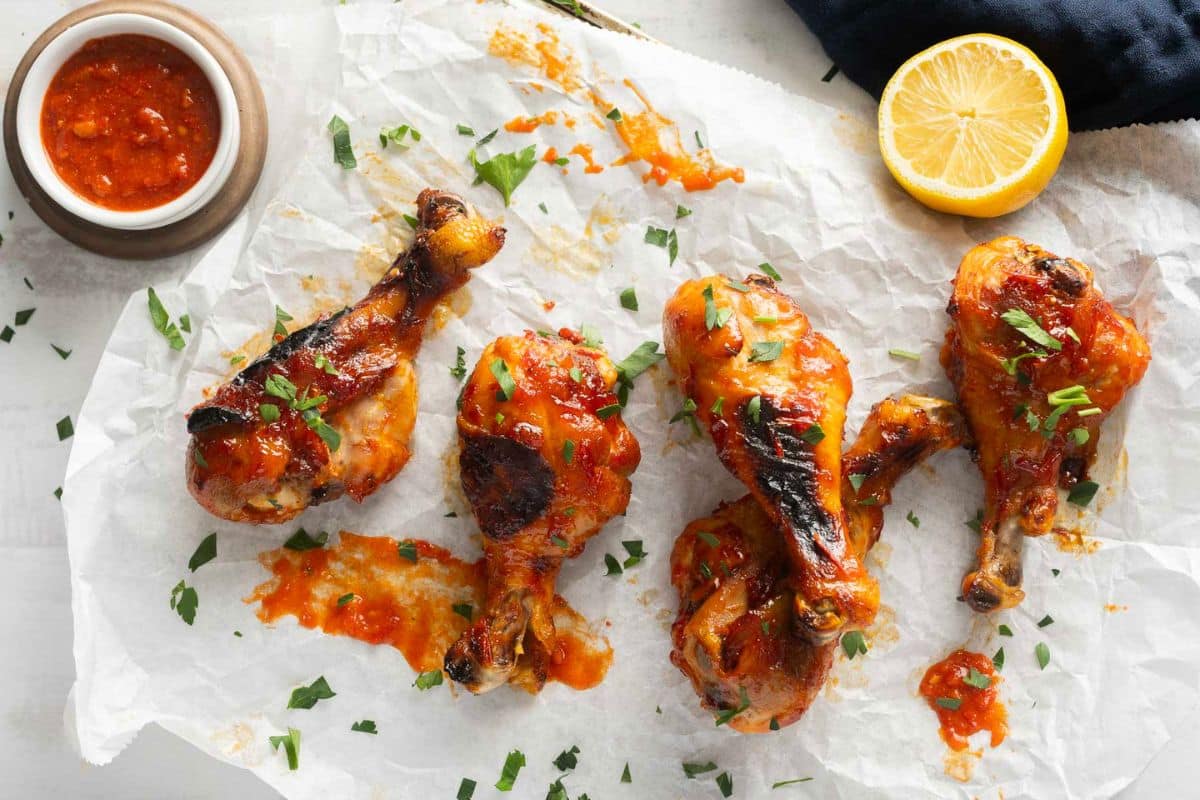 Four cooked chicken drumsticks topped with red sauce and chopped herbs on parchment paper, with a small bowl of sauce and a halved lemon-perfect for easy Family Dinners or anyone searching for quick dinner recipes.