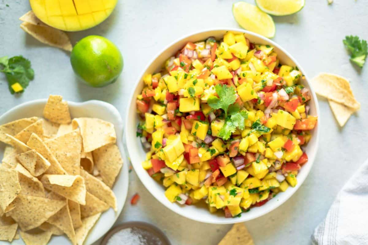 A bowl of mango salsa garnished with cilantro sits next to tortilla chips, sliced lime, and scattered cilantro leaves on a light surface-perfect for your lineup of Game Day Dips.