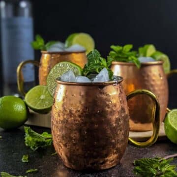Three copper mugs filled with ice and garnished with mint and lime sit on a dark surface, surrounded by fresh mint, lime halves, and beverage bottles—perfect for enjoying as refreshing autumn drinks.