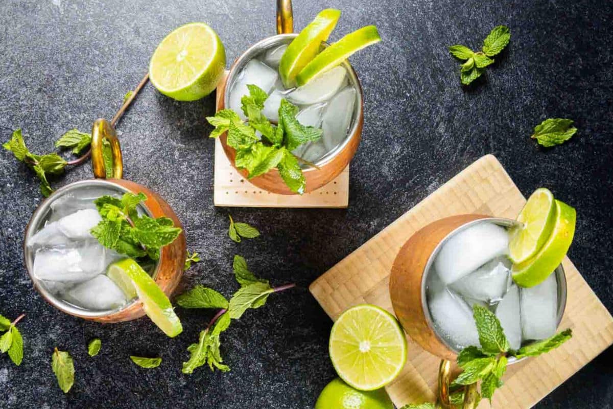 Three copper mugs filled with ice, garnished with mint sprigs and lime wedges, are placed on a dark surface-perfect for enjoying seasonal cocktails with extra lime halves and mint leaves scattered around.