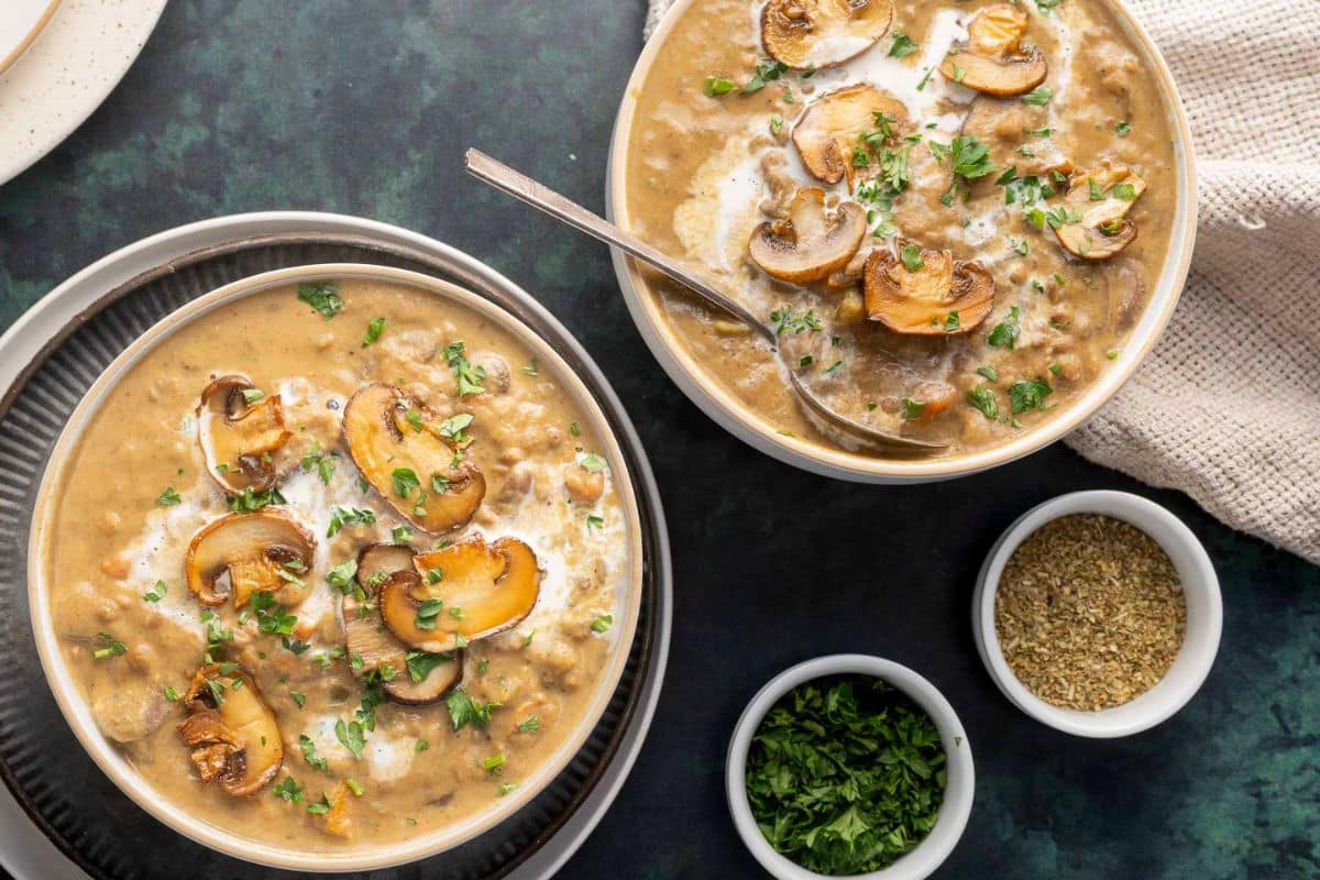 Two bowls of creamy mushroom soup, perfect for cozy soups or fall soup recipes, are topped with saut&eacute;ed mushrooms and fresh herbs, set on a dark surface beside small bowls of dried herbs and chopped parsley.