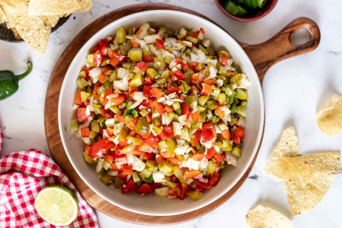 A bowl of chopped salsa made with green chiles, tomatoes, onions, and spices sits on a wooden board surrounded by tortilla chips-perfect for Game Day Dips-with jalape&ntilde;o, lime, and a red checkered napkin alongside.