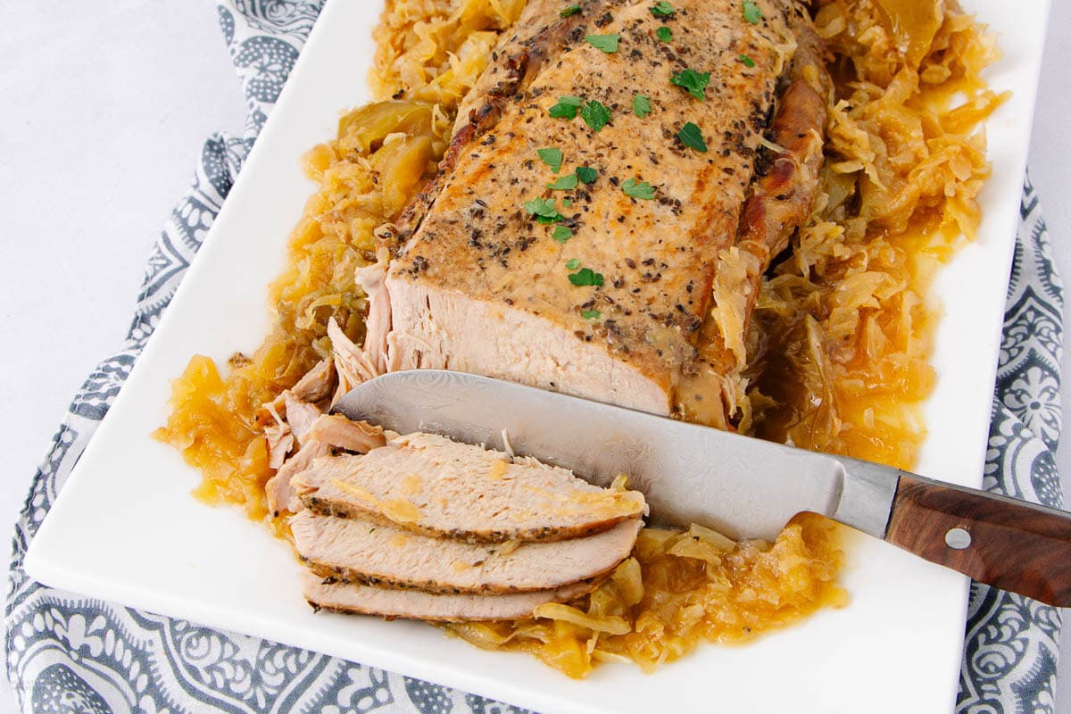 A sliced pork roast on a white platter sits atop cooked cabbage and apples, garnished with chopped parsley. Perfect for those who love seasonal apple dishes, this meal brings comforting flavors to your table. A knife rests beside the meat.