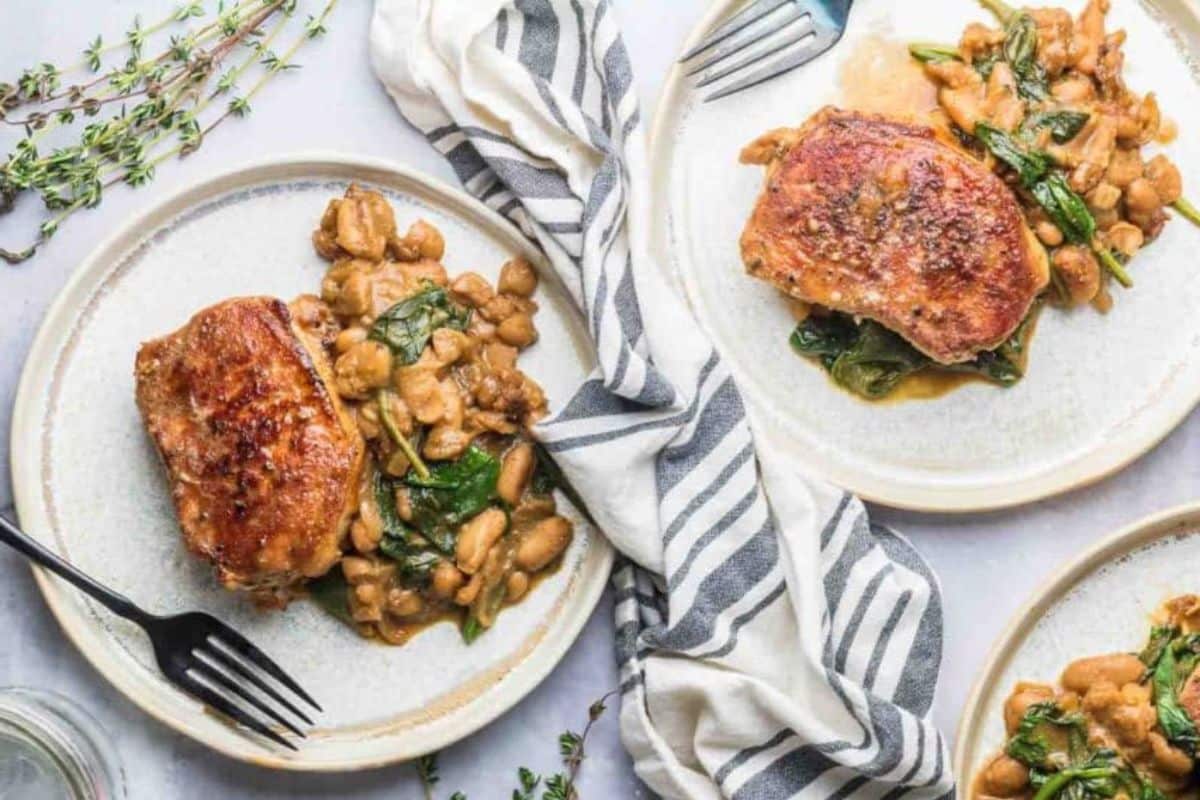 Two plates with seared pork chops, sautéed white beans, and spinach sit beside a striped napkin, garnished with fresh herbs-a perfect choice for quick dinner recipes or satisfying family dinners.