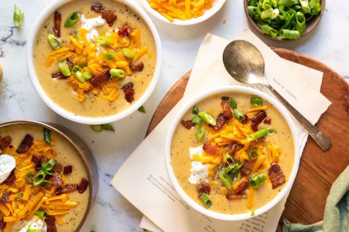 Two bowls of cozy potato soup topped with shredded cheese, bacon bits, sour cream, and chopped green onions, with a spoon and extra toppings on the side-a perfect choice for fans of creamy soups and comforting fall soup recipes.