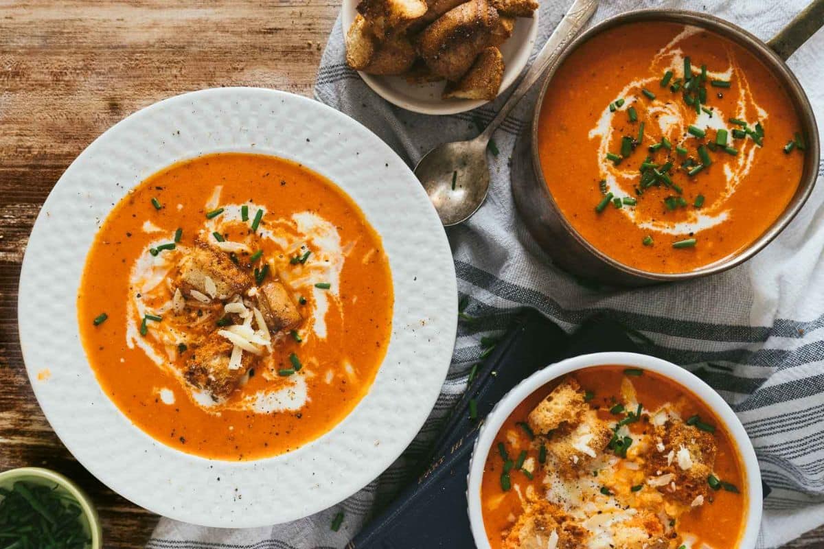 Three bowls of creamy tomato soup, one of our favorite cozy soups, garnished with cream, croutons, shredded cheese, and chives, served alongside a pot of soup and a bowl of breadsticks on a wooden table.