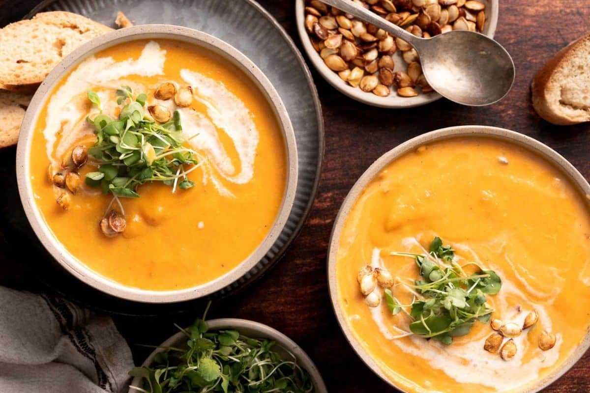 Two bowls of creamy orange soup, perfect for cozy soup recipes, topped with microgreens and roasted seeds, are served with slices of bread and a bowl of seeds on a dark wooden surface.