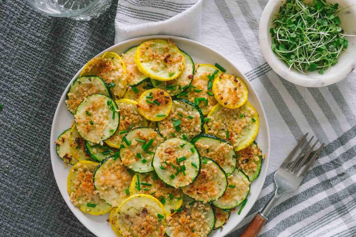 A plate of baked zucchini and yellow squash rounds topped with breadcrumbs and herbs-a delicious addition to your collection of easy squash recipes-sits beside a bowl of microgreens on a striped tablecloth.