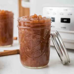 A glass jar filled with chunky applesauce sits on a counter with its lid resting beside it; another jar and cinnamon sticks are in the background.