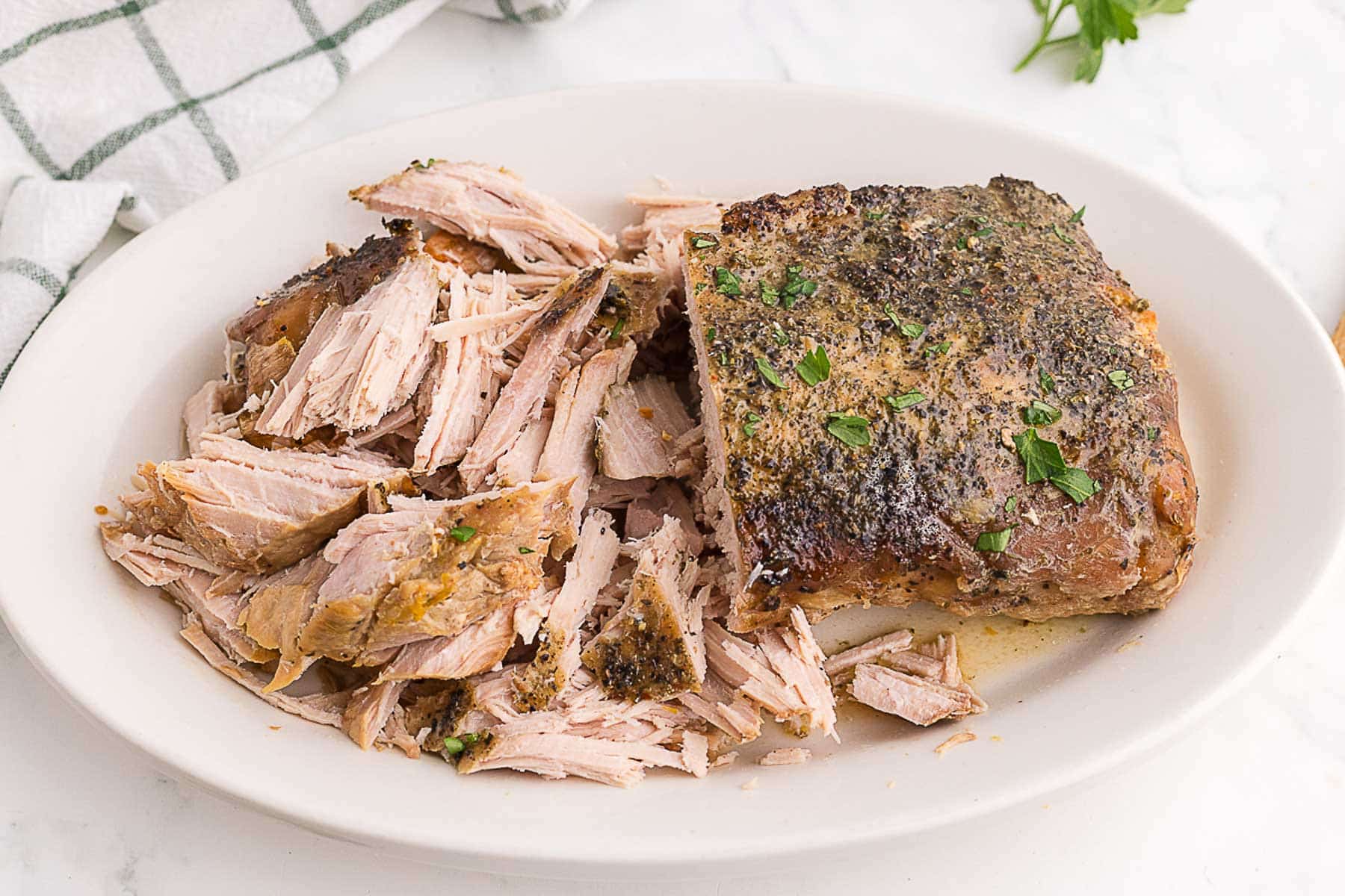 A serving platter with a partially shredded slow cooker pork roast, seasoned with herbs and garnished with chopped parsley.