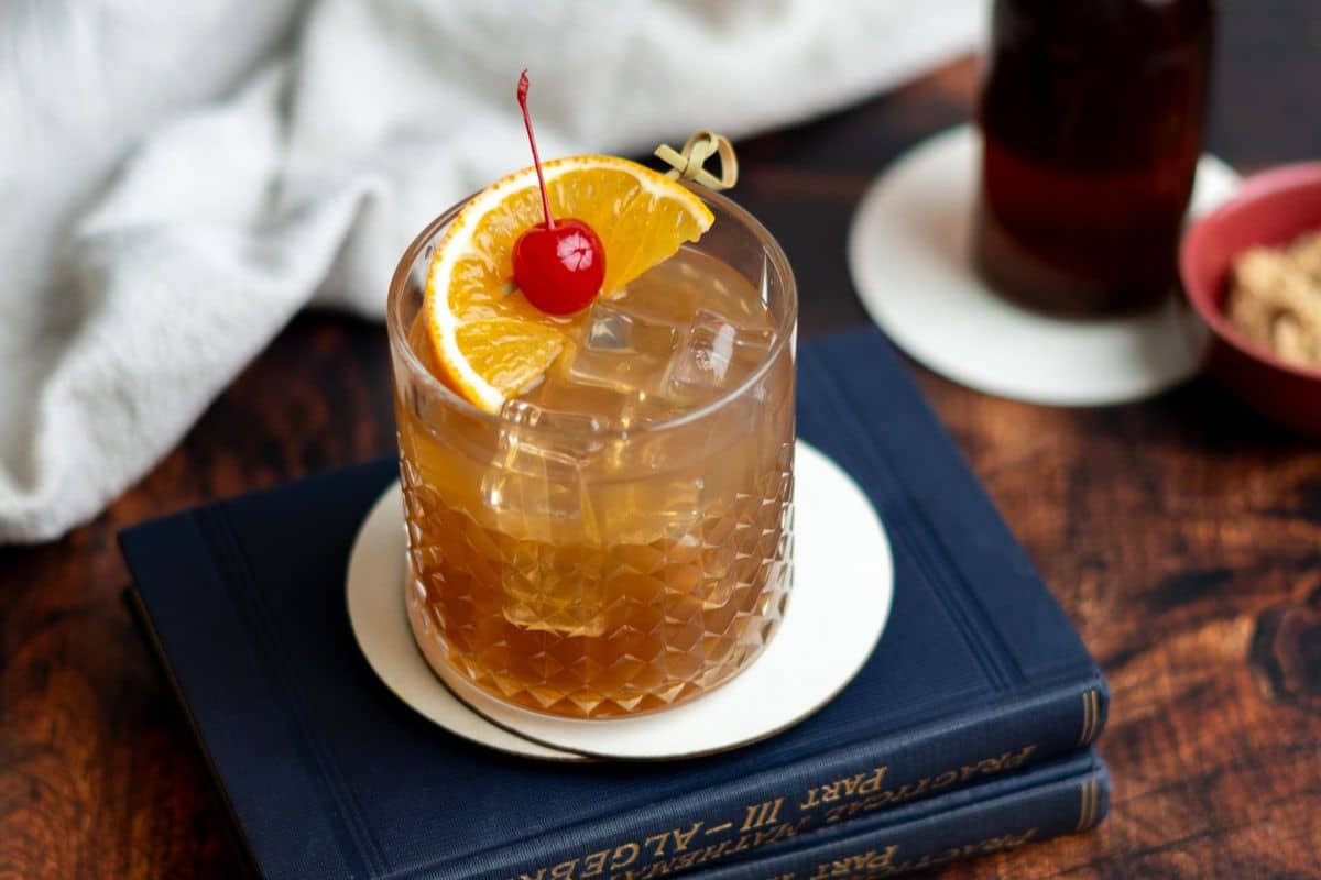 A fall cocktail with ice, garnished with an orange slice and cherry, sits on a white coaster atop two stacked blue books on a wooden table-perfect for fans of seasonal drinks.