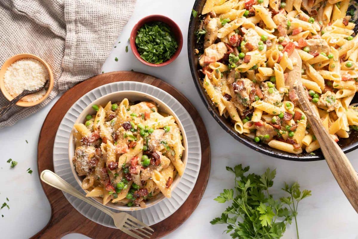 A plate and skillet of creamy penne pasta with chicken, peas, and sundried tomatoes, garnished with parsley, sit on a white surface-perfect for family dinners or as one of your favorite 30 minute meals. Grated cheese and fresh herbs complete the scene.