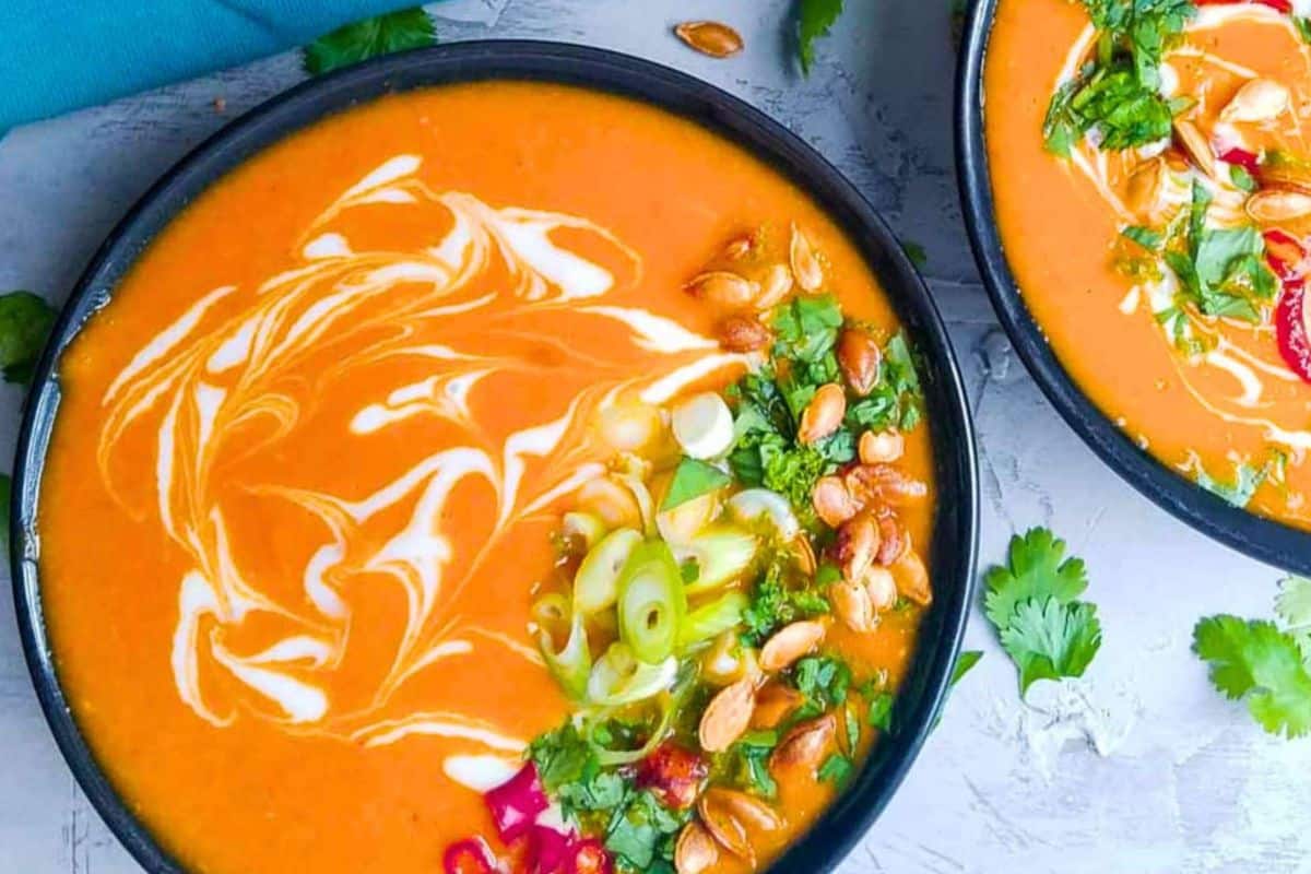 Two bowls of creamy soup, garnished with a cream swirl, chopped green onions, cilantro, peanuts, and red vegetables on a light gray surface-perfect for those seeking cozy soups or delicious fall soup recipes.