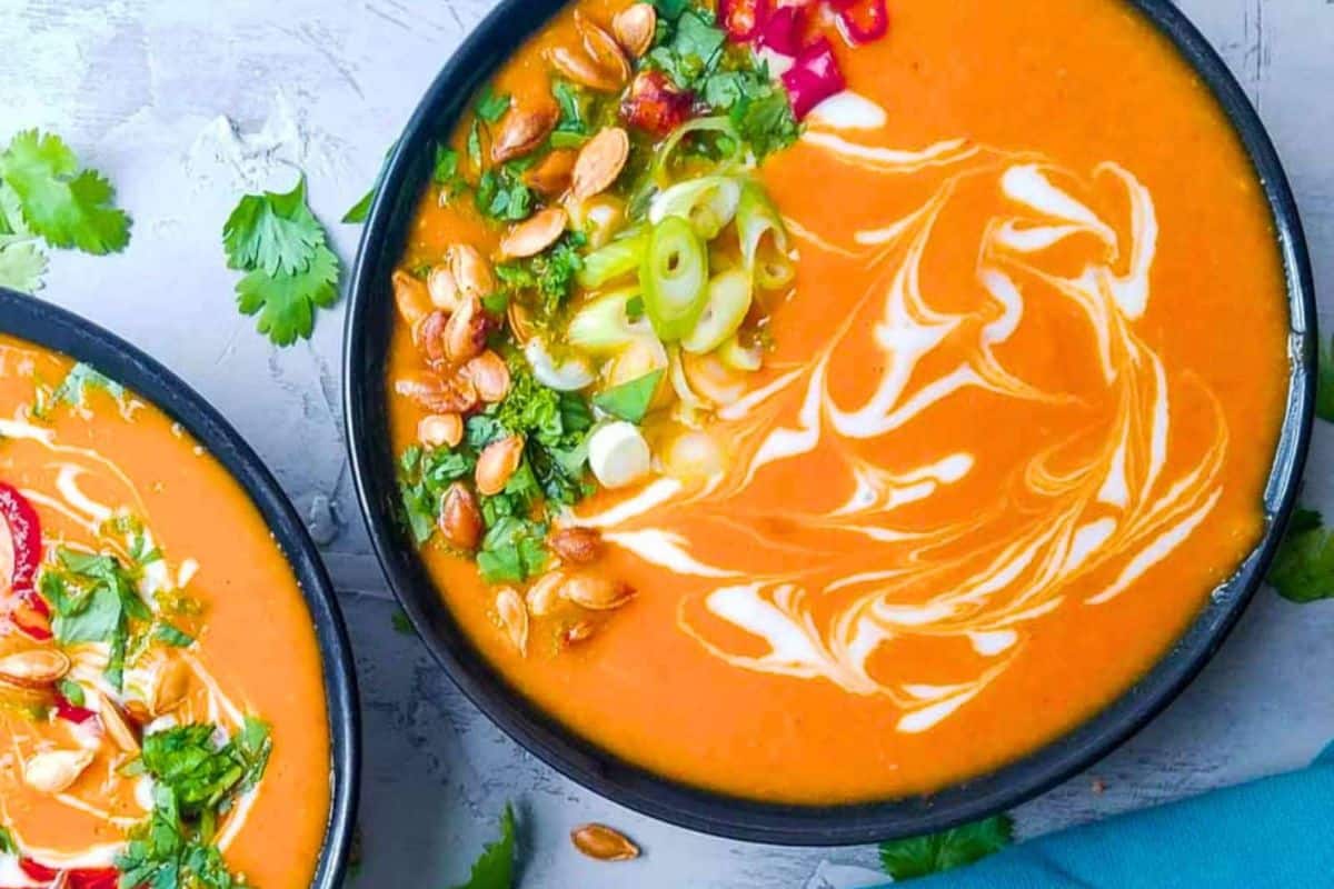 A bowl of orange soup, one of our favorite squash dishes, garnished with cream, pumpkin seeds, sliced spring onions, chopped cilantro, and red chili, with another partially visible bowl nearby.