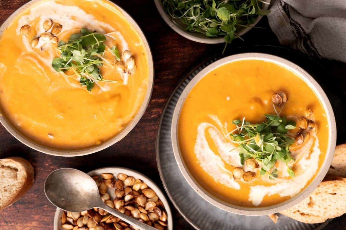 Two bowls of creamy orange soup, a delight among squash dishes, are garnished with microgreens, roasted seeds, and cream, surrounded by bread slices, a spoon, and a dish of roasted seeds.