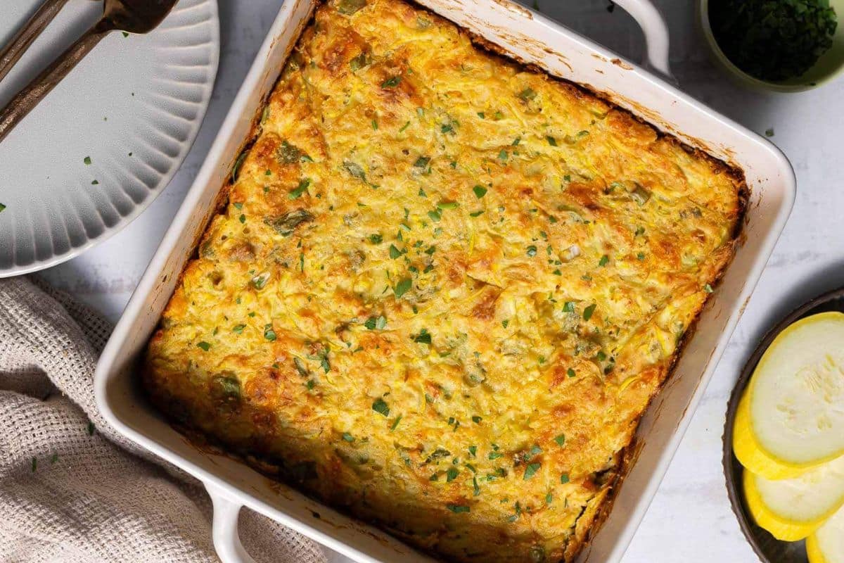A baked casserole with a golden-brown top shown in a square white dish, garnished with chopped herbs-perfect for squash recipes-next to a plate, utensils, and sliced yellow squash.