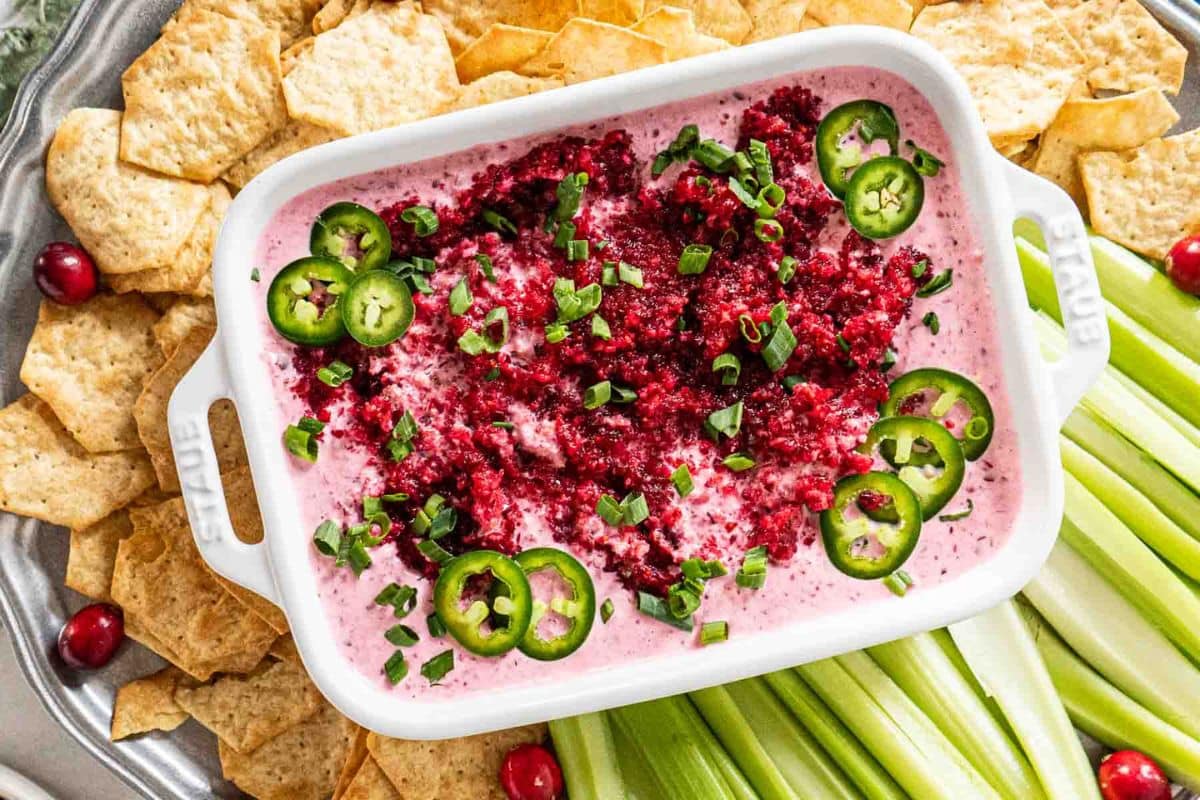 A white dish of creamy pink dip topped with chopped cranberries, sliced jalape&ntilde;os, and green onions, surrounded by tortilla chips, celery sticks, and a few cranberries-perfect for a Game Day touchdown among your favorite Game Day Dips.
