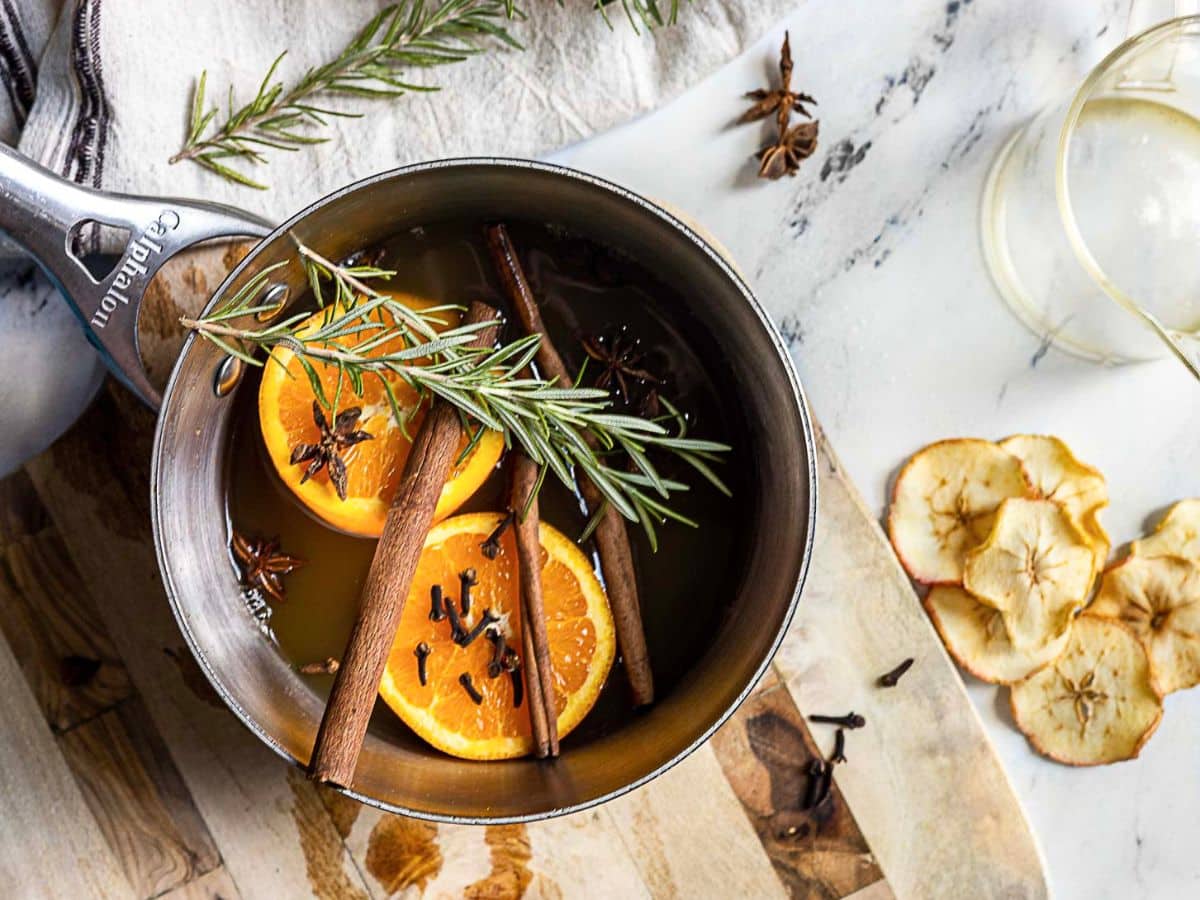 A saucepan with orange slices, cinnamon sticks, cloves, star anise, rosemary, and cider sits on a wooden board next to dried apple slices and a glass.