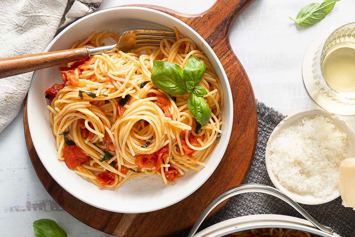 A bowl of spaghetti with tomato sauce and basil, perfect for family dinners, sits on a wooden board with a fork, grated cheese, a glass of white wine, and fresh basil leaves nearby.