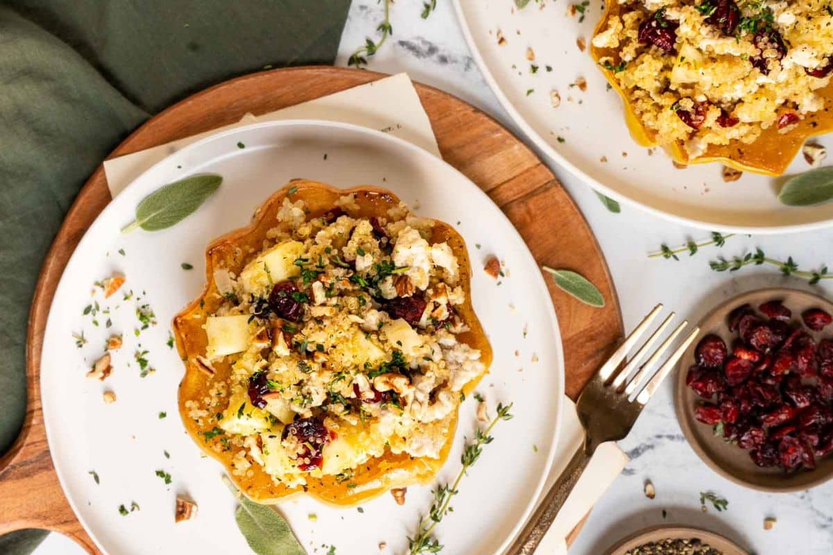 A plate with stuffed acorn squash filled with quinoa, cranberries, nuts, and herbs, garnished with fresh herbs-a delicious addition to your squash recipes-next to a fork and a small bowl of dried cranberries.