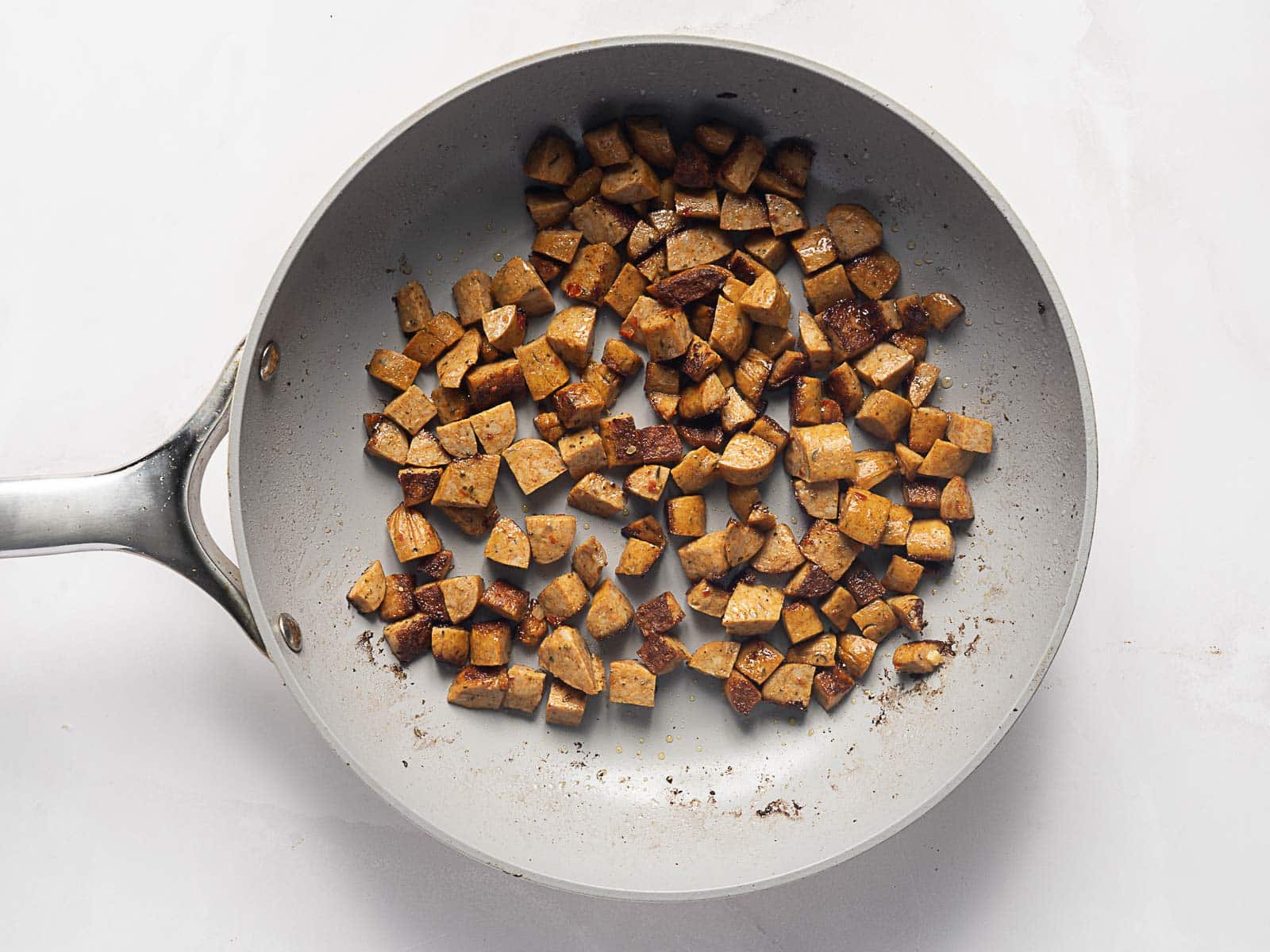 Diced tofu pieces saut&eacute;ing in a gray nonstick frying pan on a white surface.