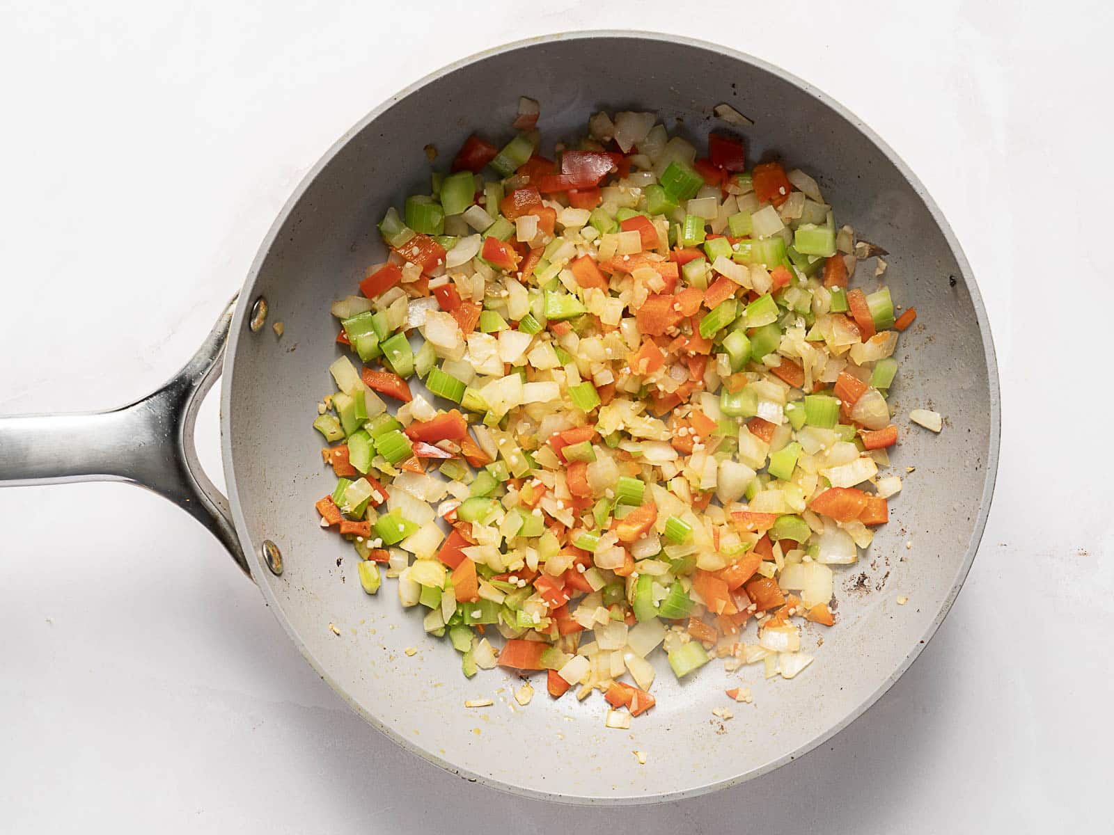 A frying pan containing chopped onions, celery, carrots, and garlic being saut&eacute;ed on a white surface.