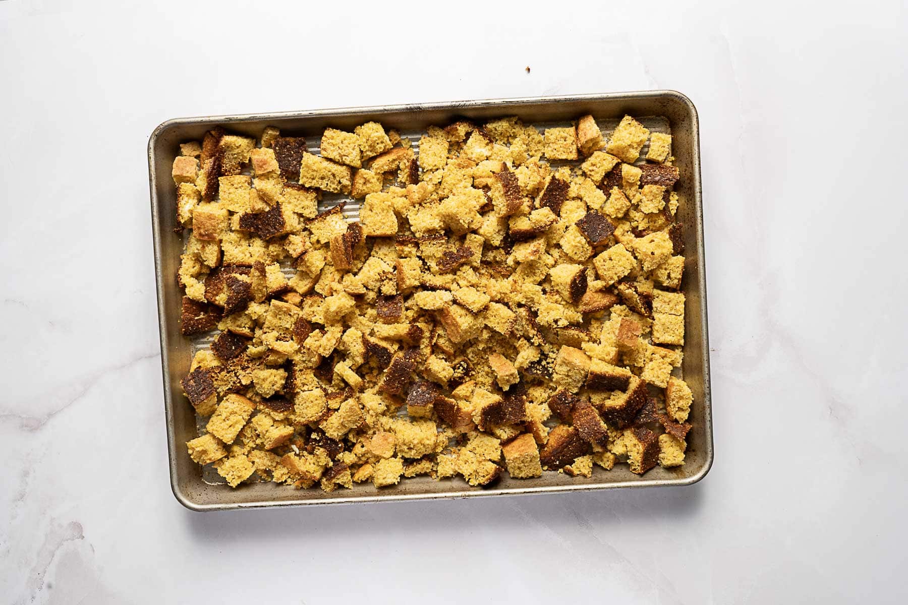 A baking sheet filled with evenly spread, golden-brown cornbread cubes on a white marble surface.
