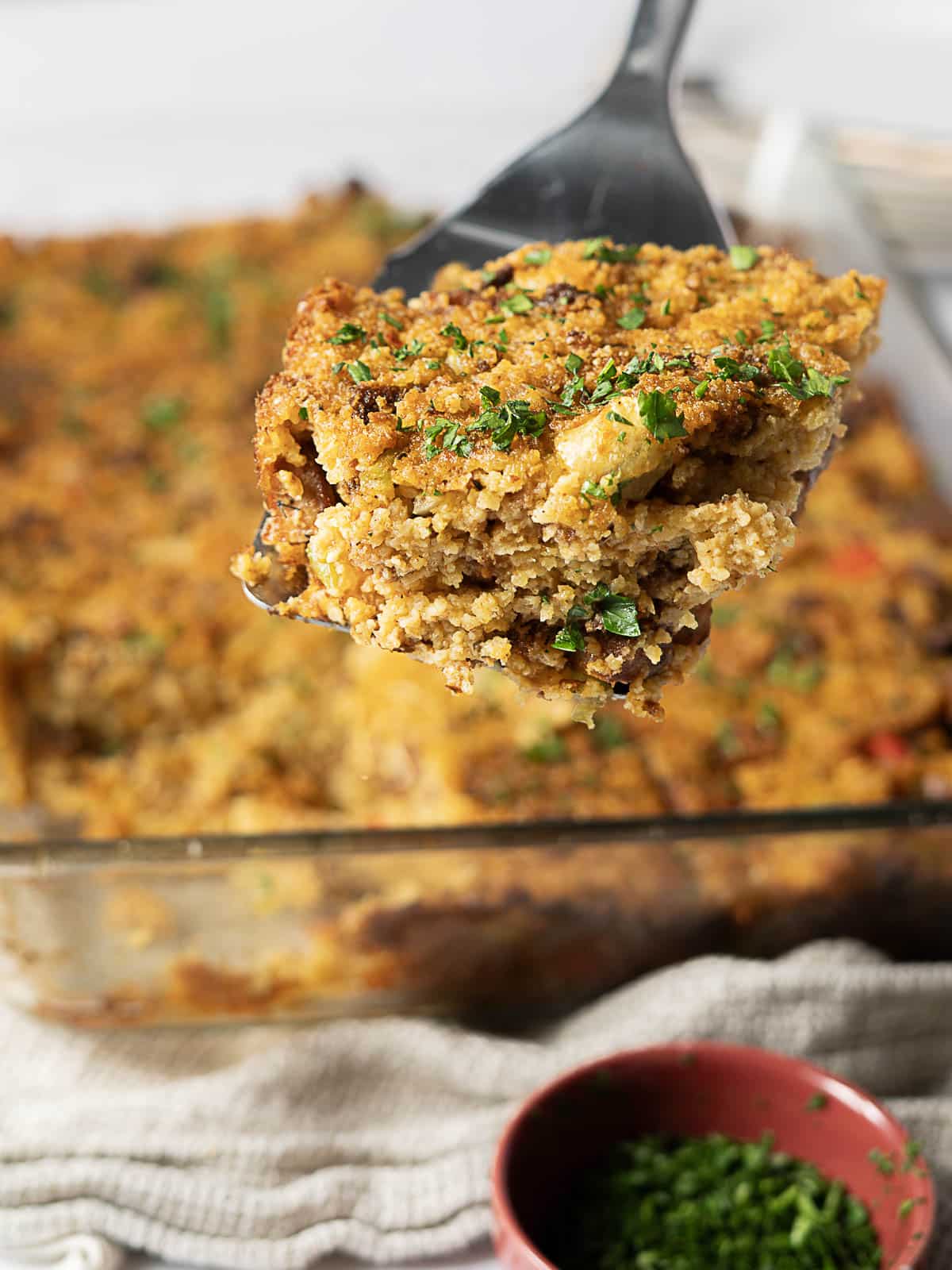 A spatula lifts a serving of baked casserole with a golden breadcrumb topping from a glass dish. A small bowl of chopped herbs sits nearby.
