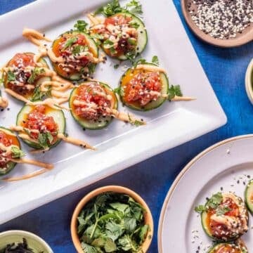 Plates of cucumber slices topped with diced vegetables, sauce, and sesame seeds make festive holiday party snacks, surrounded by bowls of herbs, sliced jalapeños, and a seed mix on a blue surface.