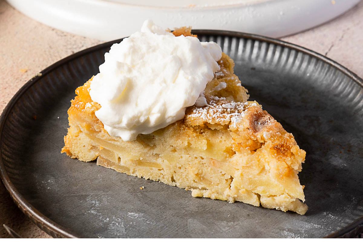 A slice of apple cake topped with a dollop of whipped cream is served on a dark plate.