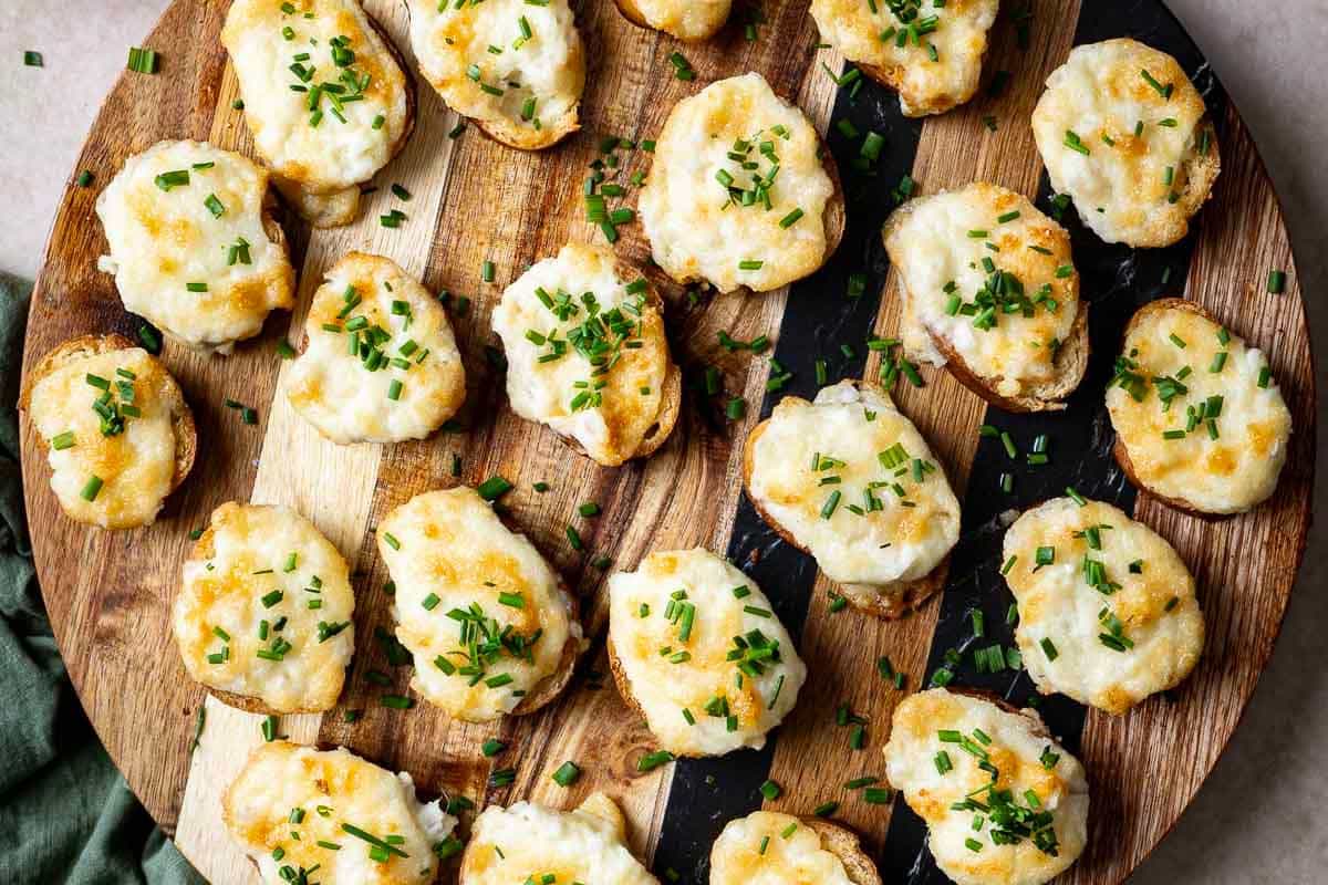 A round wooden board topped with baked potato slices covered in melted cheese and sprinkled with chopped chives-perfect for Thanksgiving appetizers or quick holiday party snacks.
