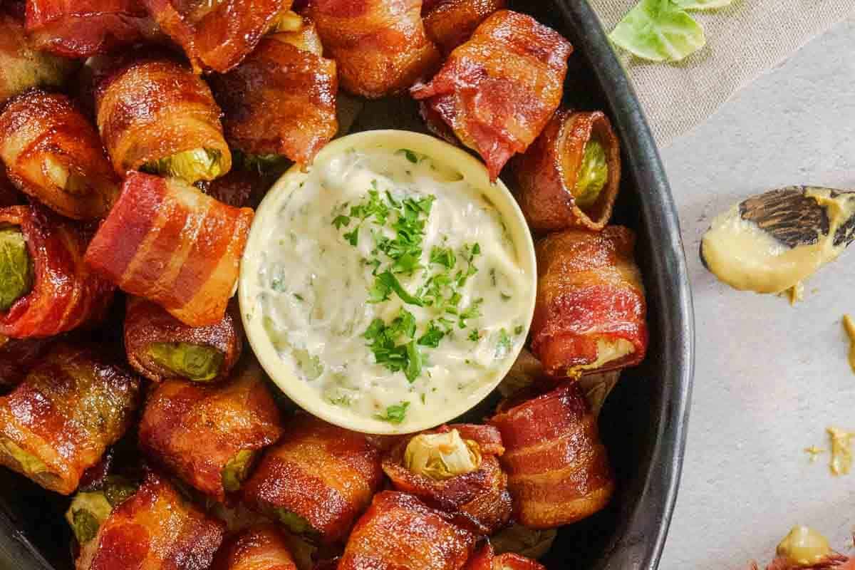Bacon-wrapped pickles arranged around a bowl of creamy dipping sauce, garnished with chopped herbs, make irresistible holiday appetizers, perfect for your next festive gathering. Served on a sleek black plate for a stunning presentation.