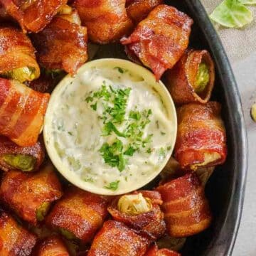 Bacon-wrapped jalapeño poppers arranged around a bowl of creamy dipping sauce, garnished with chopped herbs—perfect as Thanksgiving appetizers or for quick, last minute appetizers everyone will love.