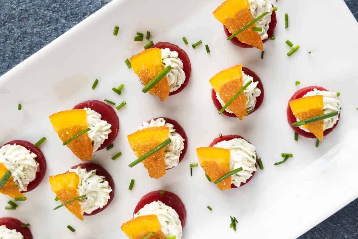 A white rectangular plate with bite-sized party appetizers topped with piped cream, orange slices, and chives, arranged neatly on a dark surface.