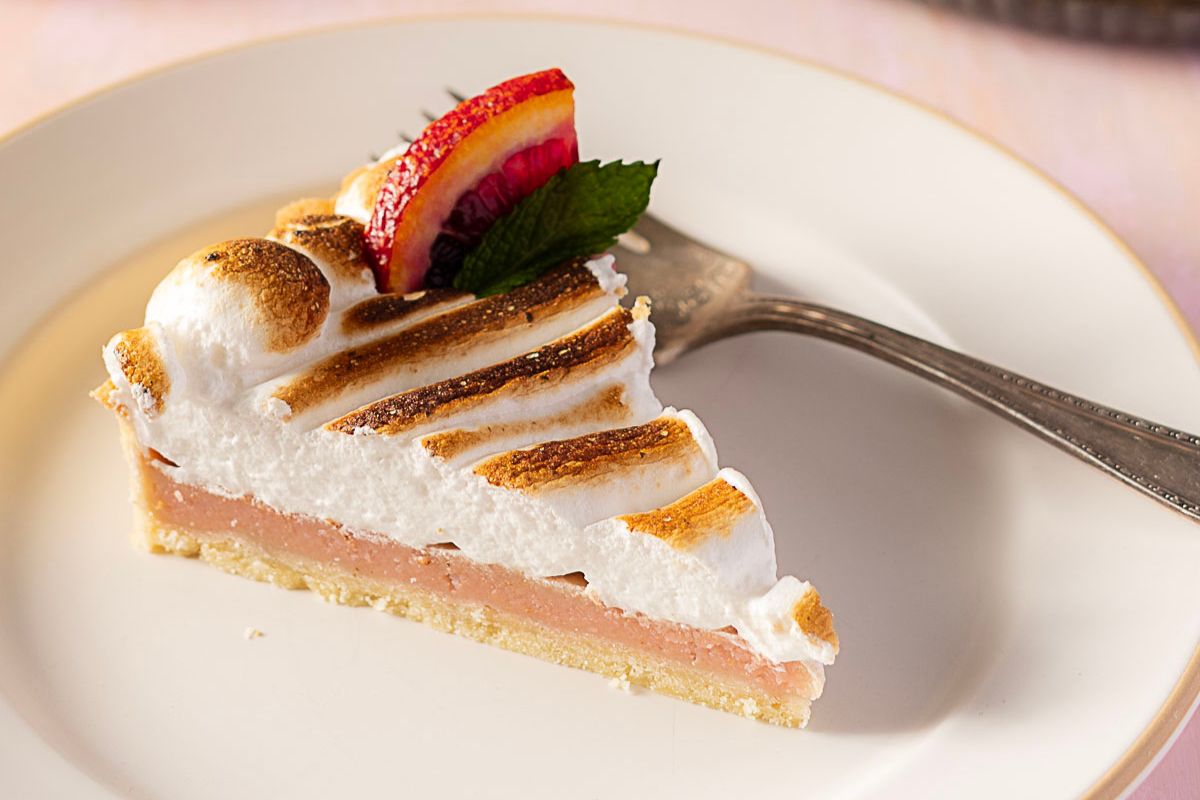 A slice of pie with a toasted meringue topping, a layer of pink filling, and a garnish of blood orange and mint on a white plate with a fork.