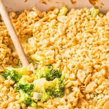 A baking dish filled with broccoli casserole topped with a golden cracker crumb crust, partially served with a wooden spoon-perfect for adding to your collection of Gluten-Free Thanksgiving recipes.