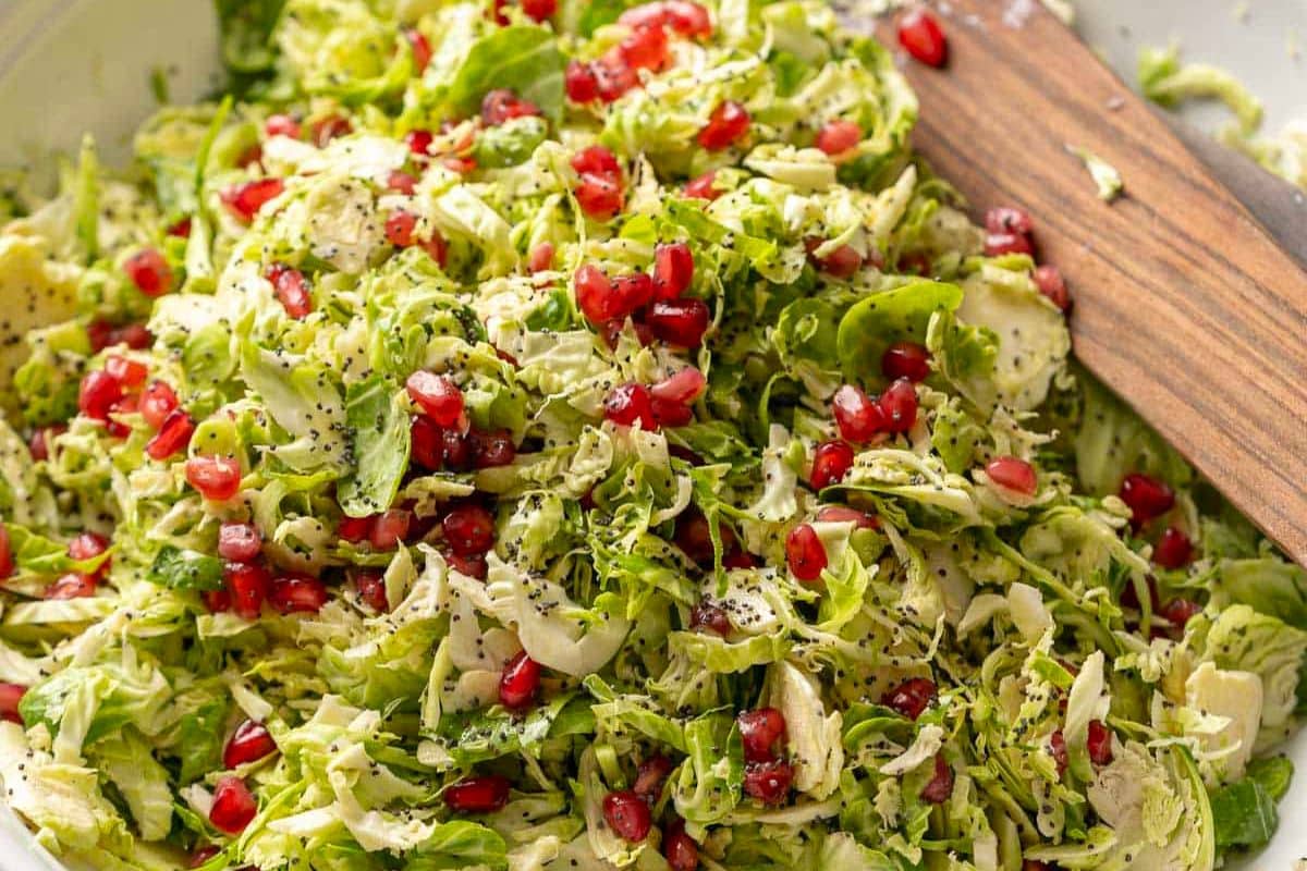 A close-up of a shredded Brussels sprout salad topped with pomegranate seeds, poppy seeds, and herbs, with a wooden serving utensil-an inspired choice for Thanksgiving sides or festive side dishes.