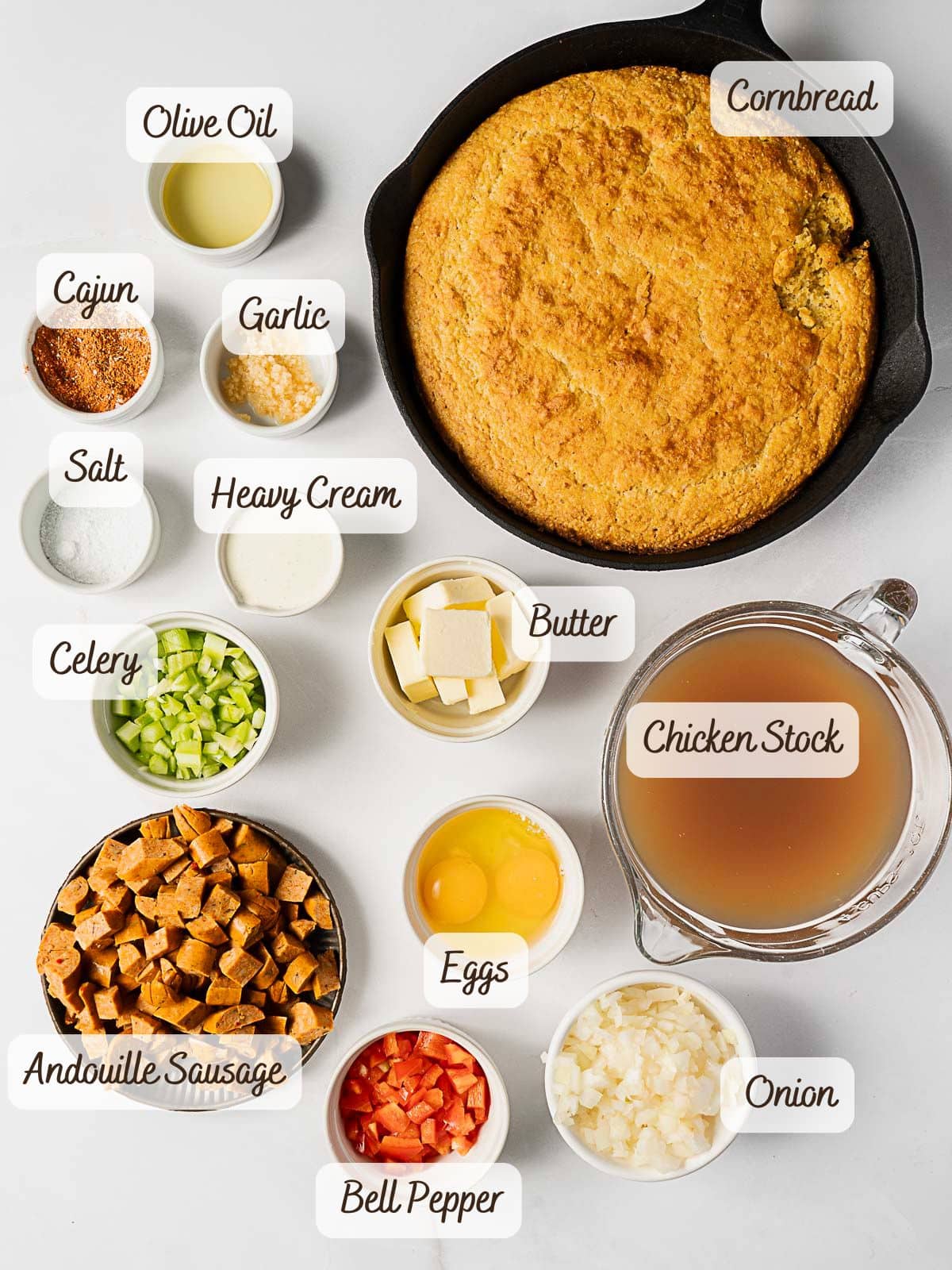 Ingredients for Cajun Cornbread Dressing arranged on a white surface, including skillet cornbread, chicken stock, and labeled bowls of vegetables, sausage, eggs, seasonings, and dairy.