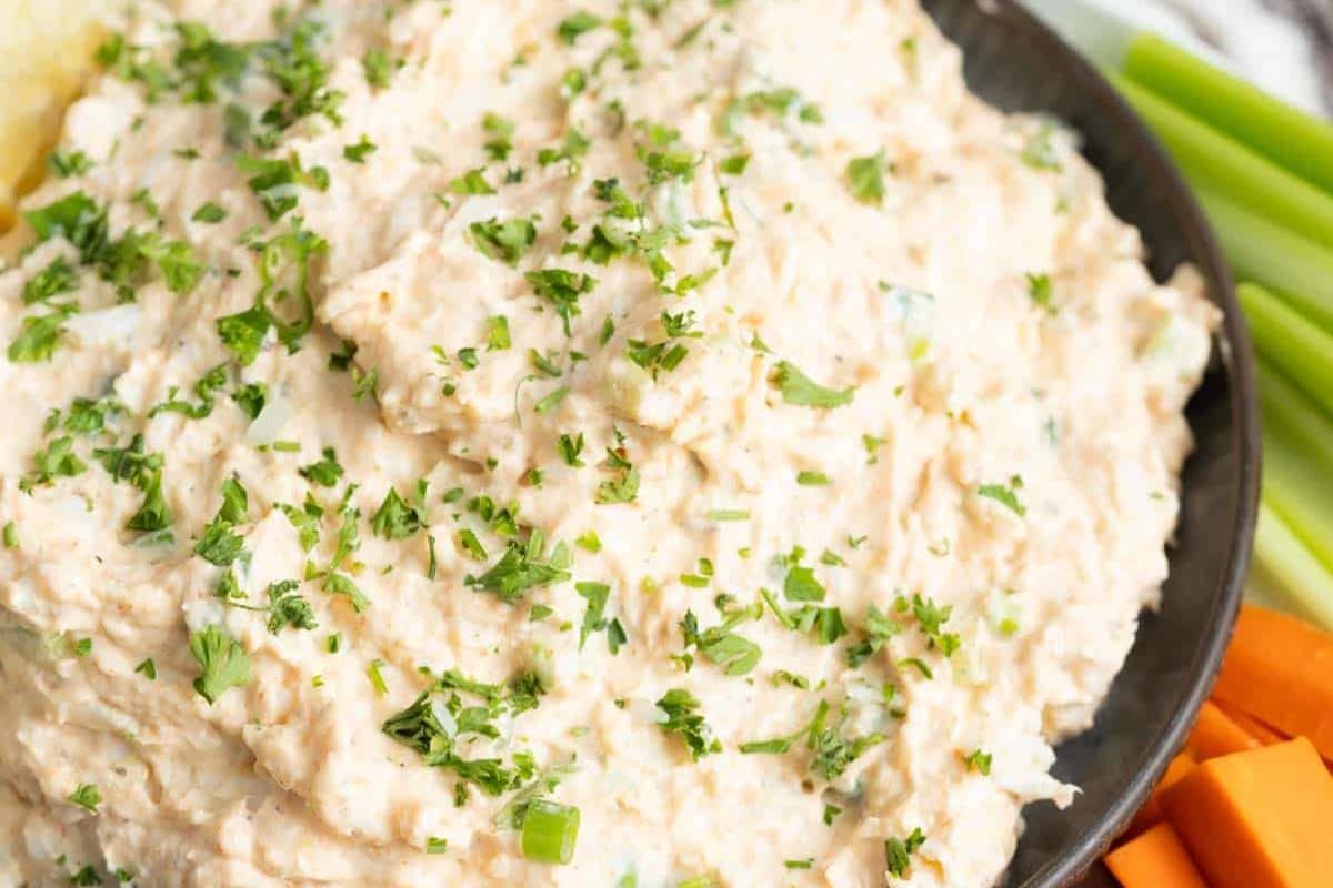 A close-up of a creamy dip topped with chopped green herbs, served in a black dish with celery and carrot sticks-perfect for holiday party food or festive party appetizers.