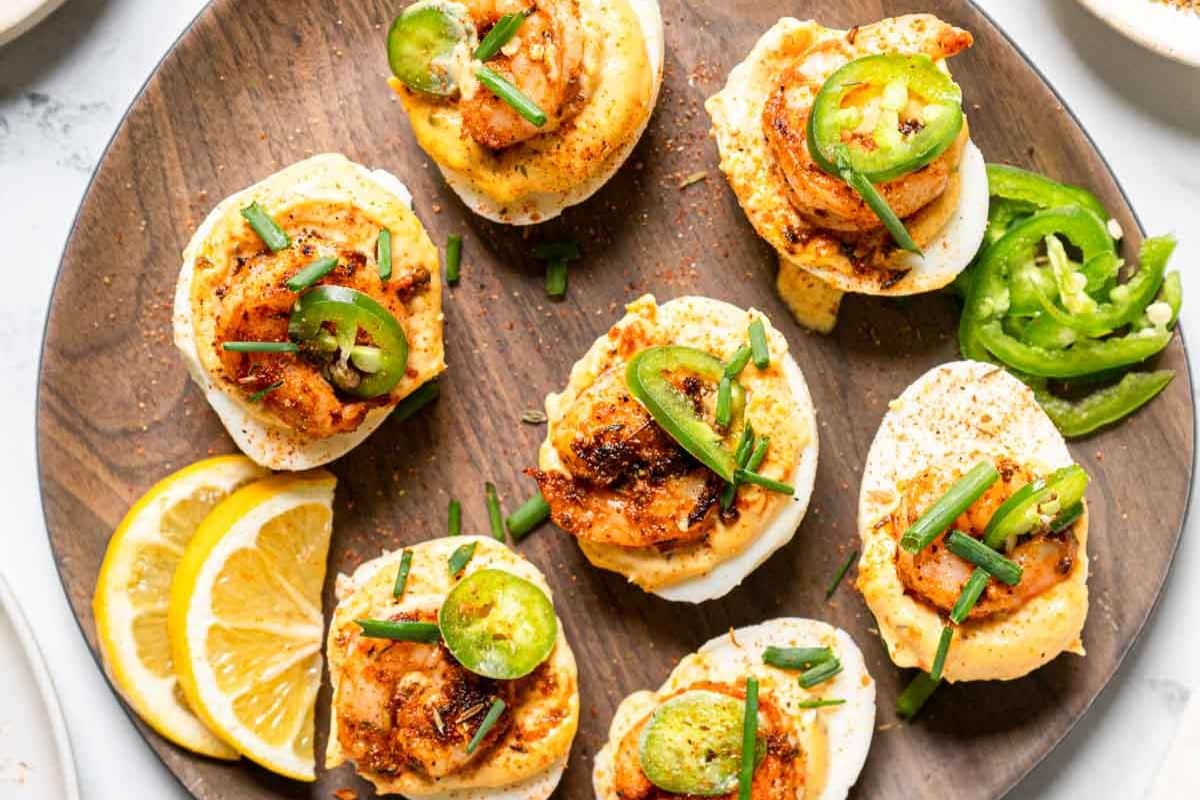 A wooden plate with deviled eggs topped with shrimp, sliced jalape&ntilde;os, chopped chives, and spices, garnished with lemon slices-perfect as festive holiday appetizers for any party.