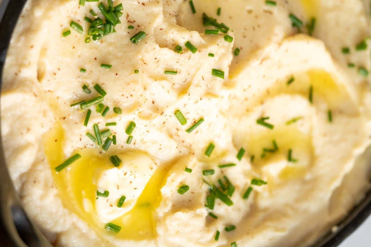 Close-up of creamy mashed potatoes topped with chopped chives, black pepper, and drizzled with melted butter-a classic choice among Thanksgiving sides.