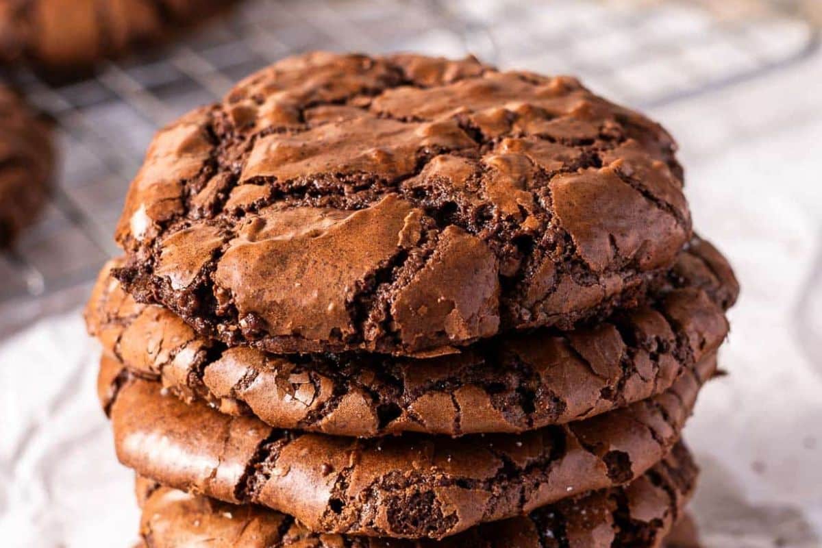 A stack of four chocolate cookies with a cracked surface sits on a sheet of parchment paper, with more cookies visible in the background on a cooling rack.