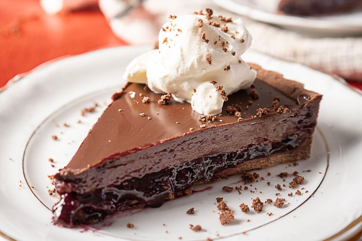 A slice of chocolate and cherry pie topped with whipped cream on a white plate.