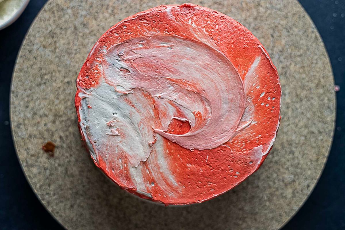 A round cake with red and white marbled frosting sits on a textured, circular surface.