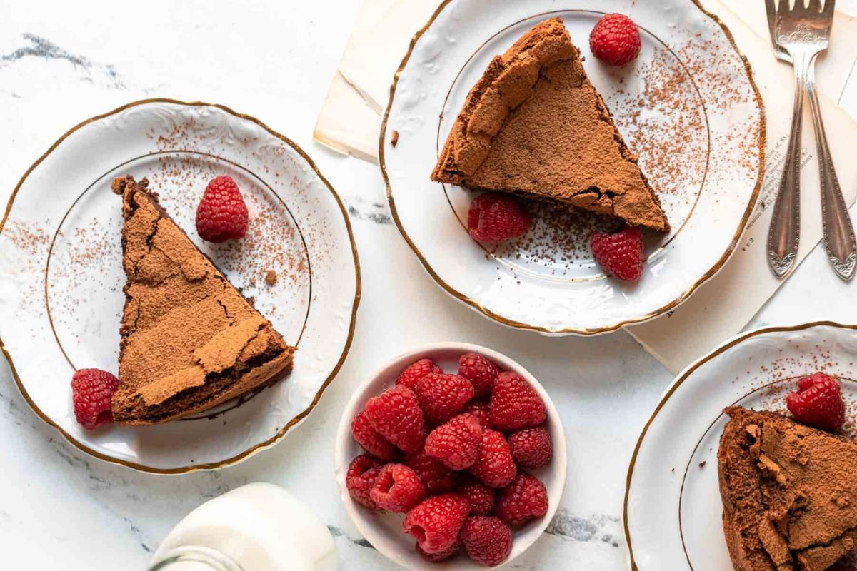 Three plates with slices of chocolate cake and raspberries, a bowl of fresh raspberries, and a bottle of milk on a marble surface.