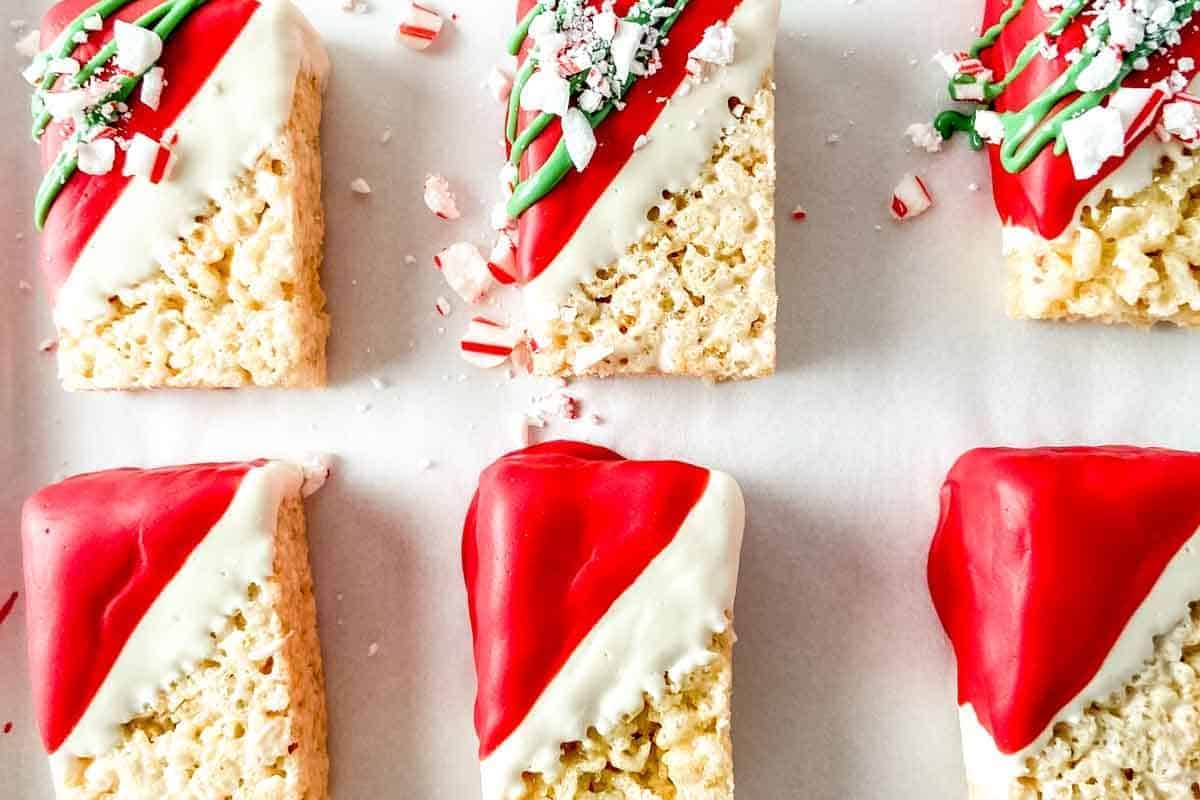 Rectangular Rice Krispies treats partially dipped in red and white chocolate, arranged on a white surface, with some topped with crushed peppermint candy and green icing.